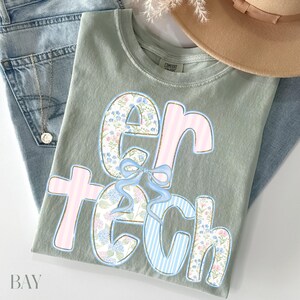 May include: Sage green t-shirt with the text "er tech" in a floral and striped design. The letters are outlined in blue and gold. A light blue bow is between the letters. The shirt is folded on top of blue jeans and a tan hat.