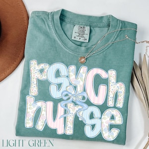 May include: Pale green t-shirt featuring the text "PSYCH NURSE" in a floral and striped design, with a light blue bow. A silver necklace with a pendant rests on the shirt. The words "LIGHT GREEN" are at the bottom.