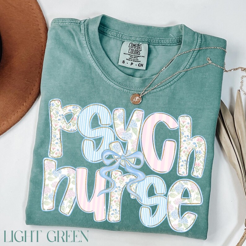 May include: Pale green t-shirt featuring the text "PSYCH NURSE" in a floral and striped design, with a light blue bow. A silver necklace with a pendant rests on the shirt. The words "LIGHT GREEN" are at the bottom.