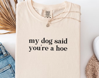 Può includere: T-shirt color crema con la scritta "my dog said you're a hoe" in nero. La maglietta è piegata e ha un'etichetta Comfort Colors. Una delicata collana con un piccolo ciondolo è appoggiata sulla maglietta. La parola "IVORY" è visibile in alto a destra.