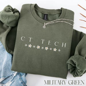 Peut inclure: Un sweat-shirt vert militaire avec le texte "CT TECH" et une rangée de marguerites blanches imprimées sur le devant.