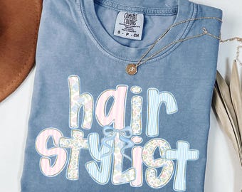 May include: A blue Comfort Colors t-shirt with the words "hair stylist" in a decorative font. The word "hair" is in pink and floral print, and "stylist" is in blue stripes and floral print. A gold necklace is draped across the shirt.