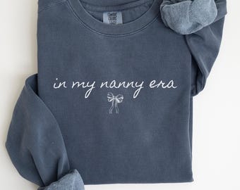 Può includere: Una felpa blu scuro con il testo "in my nanny era" in scrittura bianca con un piccolo fiocco bianco sotto. La felpa è realizzata in un materiale morbido e confortevole.