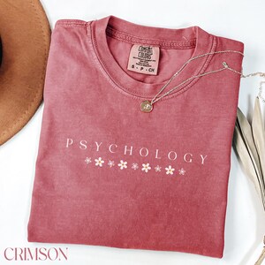 May include: Crimson red t-shirt with the text "PSYCHOLOGY" in white lettering and a row of white daisies below the text.