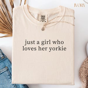 May include: Ivory-colored t-shirt with the text "just a girl who loves her yorkie." The shirt has a crew neck and short sleeves. A gold-colored necklace with a pendant is draped across the shirt. The shirt is folded on a white surface.