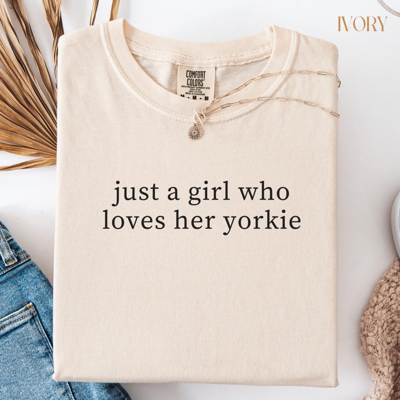 May include: Ivory-colored t-shirt with the text "just a girl who loves her yorkie." The shirt has a crew neck and short sleeves. A gold-colored necklace with a pendant is draped across the shirt. The shirt is folded on a white surface.