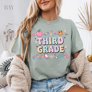 May include: A light green t-shirt with a checkered design and the text "Third Grade" in colorful letters. The shirt features a variety of school-themed graphics, including an apple, a pencil, a globe, and a smiley face.