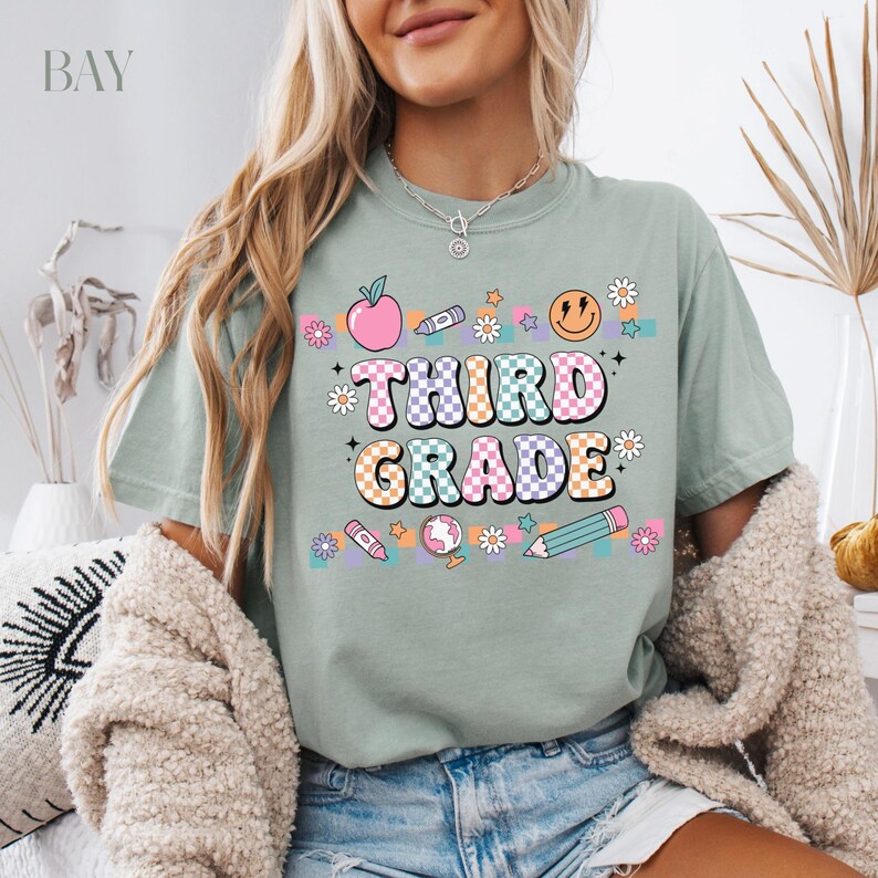May include: A light green t-shirt with a checkered design and the text "Third Grade" in colorful letters. The shirt features a variety of school-themed graphics, including an apple, a pencil, a globe, and a smiley face.