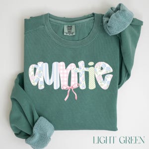 May include: Light green sweatshirt with the word "Auntie" in a whimsical font. The letters are decorated with floral and gingham patterns, and a pink bow is attached. The text "LIGHT GREEN" is at the bottom.