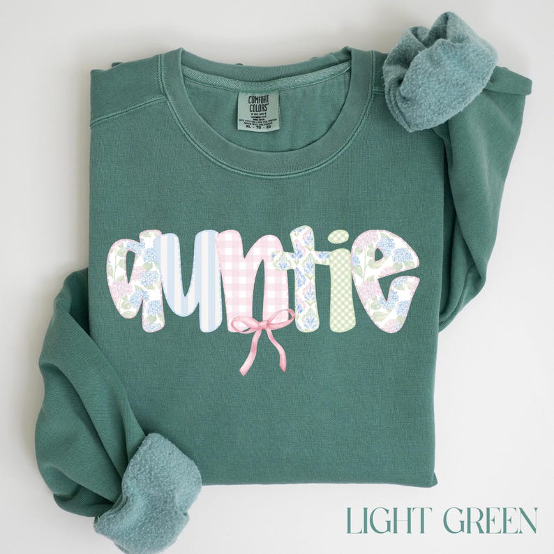 May include: Light green sweatshirt with the word "Auntie" in a whimsical font. The letters are decorated with floral and gingham patterns, and a pink bow is attached. The text "LIGHT GREEN" is at the bottom.