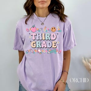 May include: A lavender t-shirt with the text "Third Grade" in colorful, checkered letters. The shirt is decorated with a variety of school-themed graphics, including an apple, a pencil, a crayon, a smiley face, and a globe.