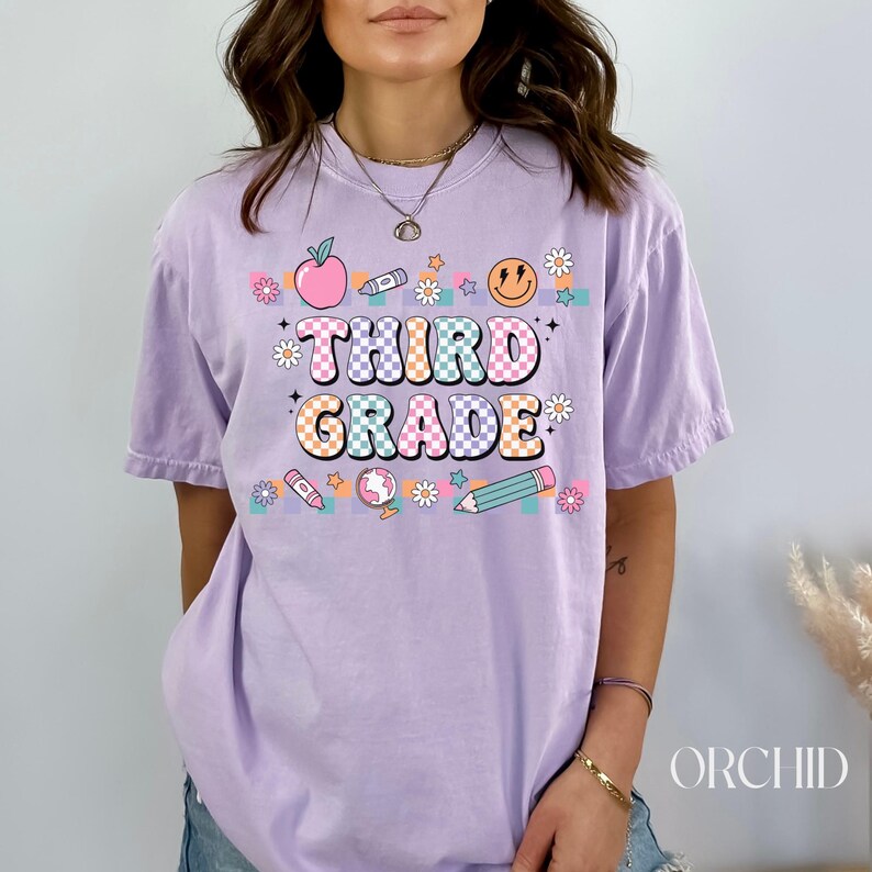 May include: A lavender t-shirt with the text "Third Grade" in colorful, checkered letters. The shirt is decorated with a variety of school-themed graphics, including an apple, a pencil, a crayon, a smiley face, and a globe.