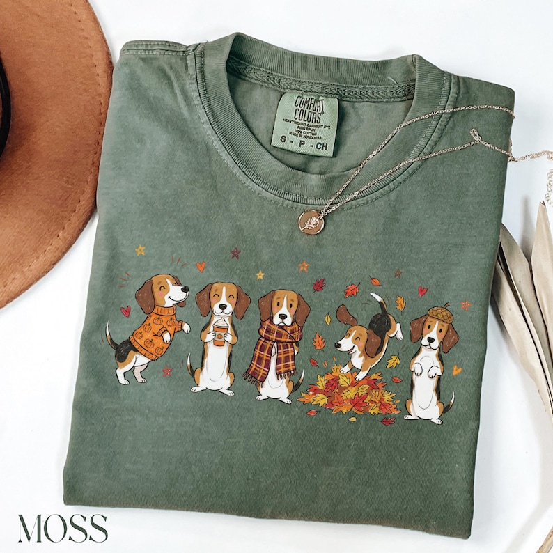 Puede incluir: Una camiseta de manga corta color verde musgo con un gr&aacute;fico de cinco perros beagle con ropa de oto&ntilde;o, incluyendo su&eacute;teres, bufandas y sombreros, rodeados de hojas. La etiqueta interior del cuello dice Comfort Colors, 100% algod&oacute;n, S-P-CH.