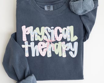 May include: A denim blue sweatshirt with the words "Physical Therapy" in a decorative font. The letters are patterned with pink gingham, blue floral, and green checkered designs, tied with a pink bow. The word "DENIM" is at the bottom.