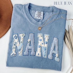 Peut inclure: Un sweat-shirt bleu clair Comfort Colors avec le mot "NANA" brodé sur le devant dans un motif patchwork. Les lettres présentent des motifs floraux, à pois et à carreaux. Un collier en or avec un pendentif repose sur le chemisier.