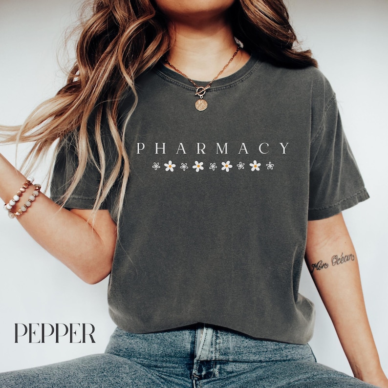 May include: A dark gray t-shirt with the word "PHARMACY" printed in white letters. The shirt has a row of small white flowers printed below the word. The model is wearing the shirt with blue jeans.