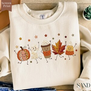 May include: A cream-coloured crewneck sweatshirt featuring a screen-printed graphic of five smiling autumn-themed characters: a pumpkin, a ghost, a coffee cup, a maple leaf, and a piece of candy corn, all surrounded by small scattered stars.
