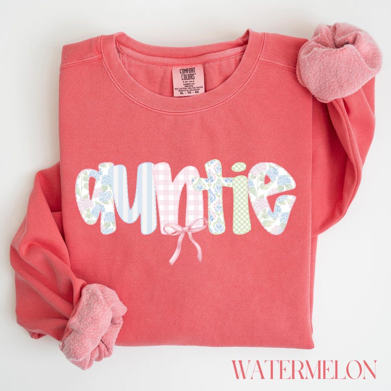 May include: Coral-colored sweatshirt with the word "AUNTIE" in a whimsical font. The letters feature various patterns, including stripes, gingham, and floral designs. A small pink bow is placed under the letters. The word "WATERMELON" is printed at the bottom.