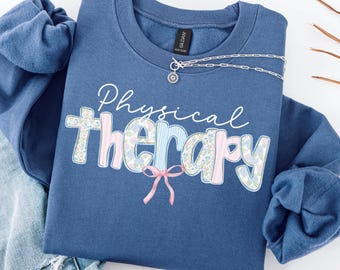 May include: Indigo blue sweatshirt with the words "Physical Therapy" in white script and floral and striped applique. A silver necklace with a small pendant is draped across the top. The bottom of the sweatshirt is folded over a pair of blue jeans.