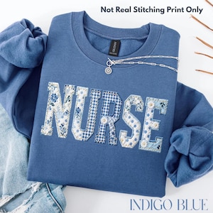 May include: Indigo blue sweatshirt featuring the word "NURSE" in a patchwork design with floral, polka dot, and gingham patterns. A silver necklace with a pendant rests on the sweatshirt. The text "Not Real Stitching Print Only" is at the top.