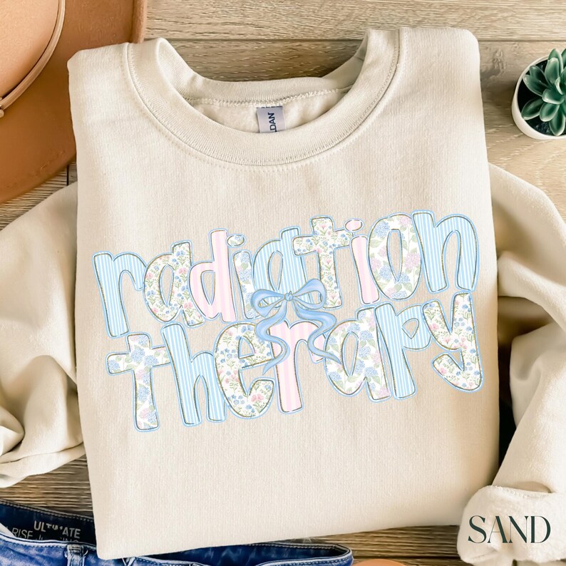 May include: A cream-colored sweatshirt with the words "radiation therapy" in a light blue and pink design. The word "radiation" is in a striped pattern, and "therapy" is in a floral pattern. A light blue bow is between the words.