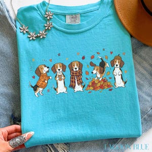 Puede incluir: Una camiseta de manga corta de color turquesa brillante con un estampado gr&aacute;fico de cinco perros beagle con ropa de oto&ntilde;o, incluyendo jers&eacute;is y bufandas, jugando entre hojas ca&iacute;das. La camiseta tiene una etiqueta de la marca Comfort Colors.