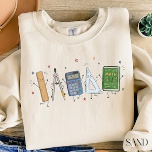 May include: A cream-colored crewneck sweatshirt featuring a graphic print of five anthropomorphic school supplies: a wooden ruler, a metal compass, a blue calculator, a clear triangle ruler, and a green book labeled MATH with small stars.