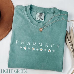 May include: Light green t-shirt with the word "PHARMACY" in white lettering and a row of white daisies below. The shirt has a tag that says "COMFORT COLORS" and "S - P - CH".