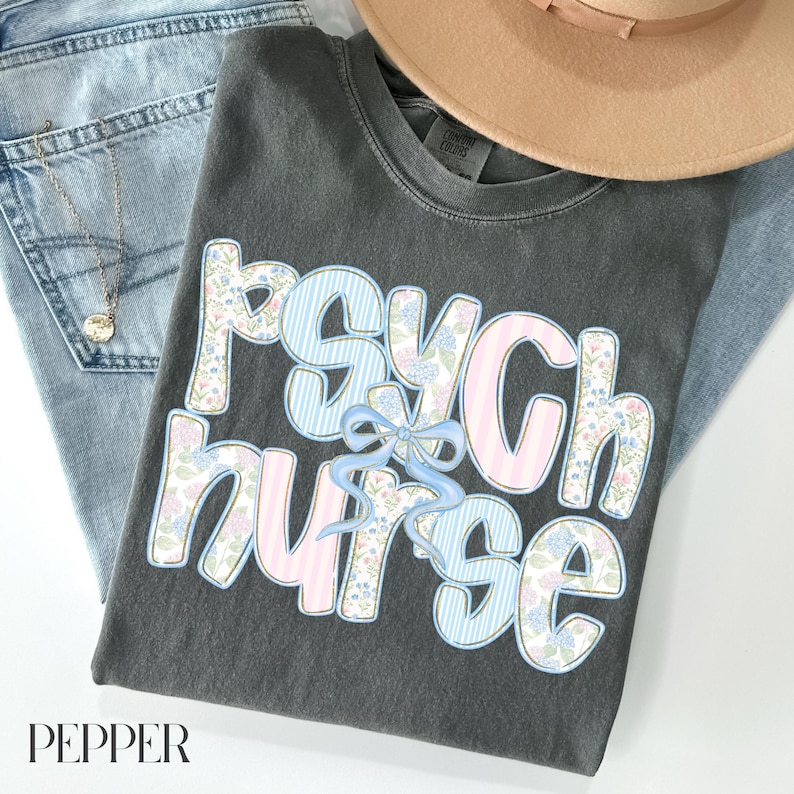 May include: A dark grey t-shirt with the words "Psych Nurse" in a floral and striped design. The shirt is paired with light wash jeans, a gold necklace, and a beige hat. The word "PEPPER" is at the bottom.