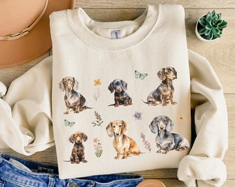 May include: A cream-colored sweatshirt with a graphic print of six dachshund dogs in various poses. The dogs are surrounded by flowers and butterflies. The sweatshirt is laid out on a wooden surface with a pair of jeans and a hat.