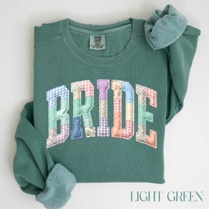 May include: Light green sweatshirt with the word "BRIDE" appliquéd across the front in a patchwork design. The patchwork letters feature various patterns and colours, including floral, gingham, and solid fabrics. The sweatshirt has a crew neck and long sleeves.
