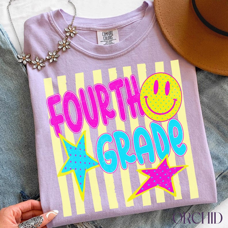 Op de afbeelding: Een lila T-shirt met de tekst "FOURTH GRADE" in roze en blauw, versierd met een gele smiley en sterren. Het shirt heeft een ronde hals en is gemaakt van een zacht materiaal.