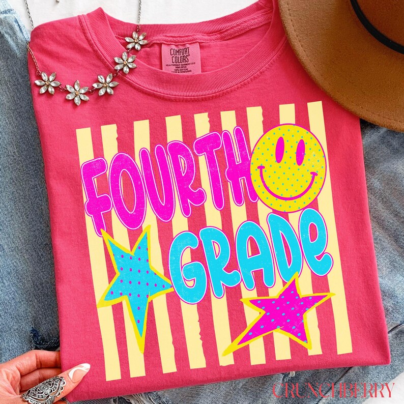 Op de afbeelding: Een roze t-shirt met de woorden "FOURTH GRADE" in een speels lettertype, geaccentueerd met een gele smiley en sterren. Het ontwerp heeft gele verticale strepen. Een ketting en een hoed zijn zichtbaar op de achtergrond.