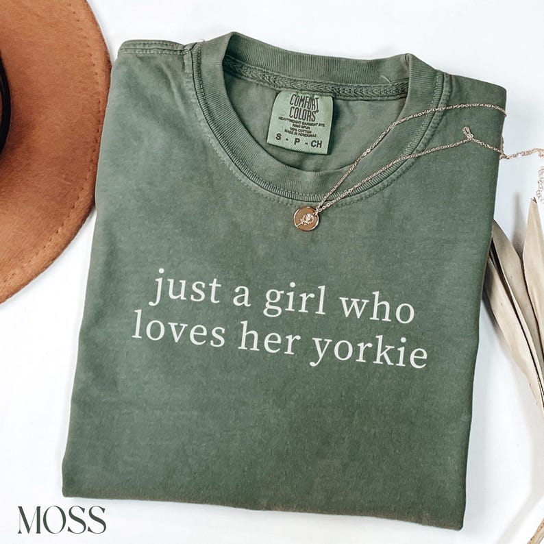May include: An olive green t-shirt with the text "just a girl who loves her yorkie" in white. A gold necklace with a pendant rests on the shirt. The shirt has a Comfort Colors tag.