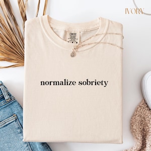 May include: Ivory t-shirt with the text "normalize sobriety" printed in black.