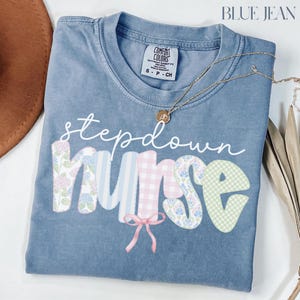 May include: A blue Comfort Colors t-shirt with the words "stepdown nurse" in a whimsical font. The letters are decorated with floral, striped, and gingham patterns. A gold necklace with a pendant is draped across the shirt.