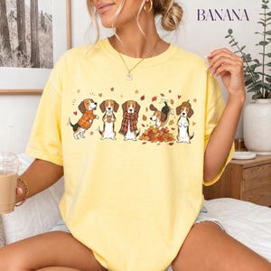Puede incluir: Una camiseta de manga corta de color amarillo p&aacute;lido con un estampado gr&aacute;fico de cinco perros beagle en varias poses oto&ntilde;ales, incluyendo con un su&eacute;ter de calabazas, una bufanda de cuadros, jugando entre hojas y con un gorro de bellota, con la palabra BANANA.