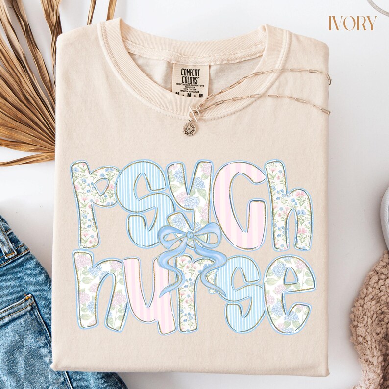 May include: Cream-coloured t-shirt featuring the words "Psych Nurse" in a playful font. The letters are adorned with floral and striped patterns in pastel blue and pink, with a light blue bow. A delicate gold necklace is draped across the shirt.