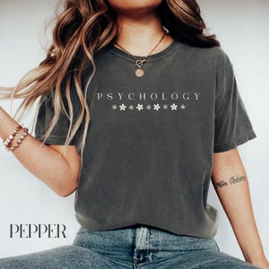 May include: A dark gray t-shirt with the text "PSYCHOLOGY" printed in white. The text is surrounded by a row of small white flowers.