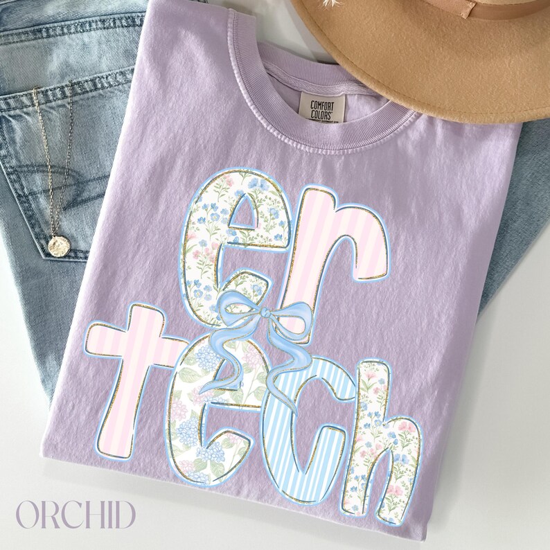 May include: Orchid-colored t-shirt with the text "er tech" in a decorative font. The letters feature floral, striped, and solid patterns with a light blue bow. A pair of blue jeans and a necklace are visible.