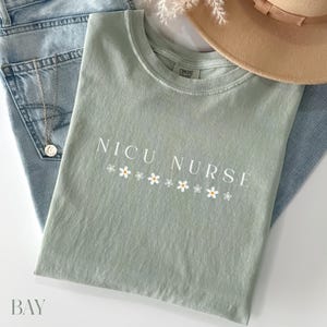 Peut inclure: Un t-shirt vert clair avec le texte "NICU NURSE" en lettres blanches et une rang&eacute;e de petites fleurs blanches avec des centres jaunes sous le texte.