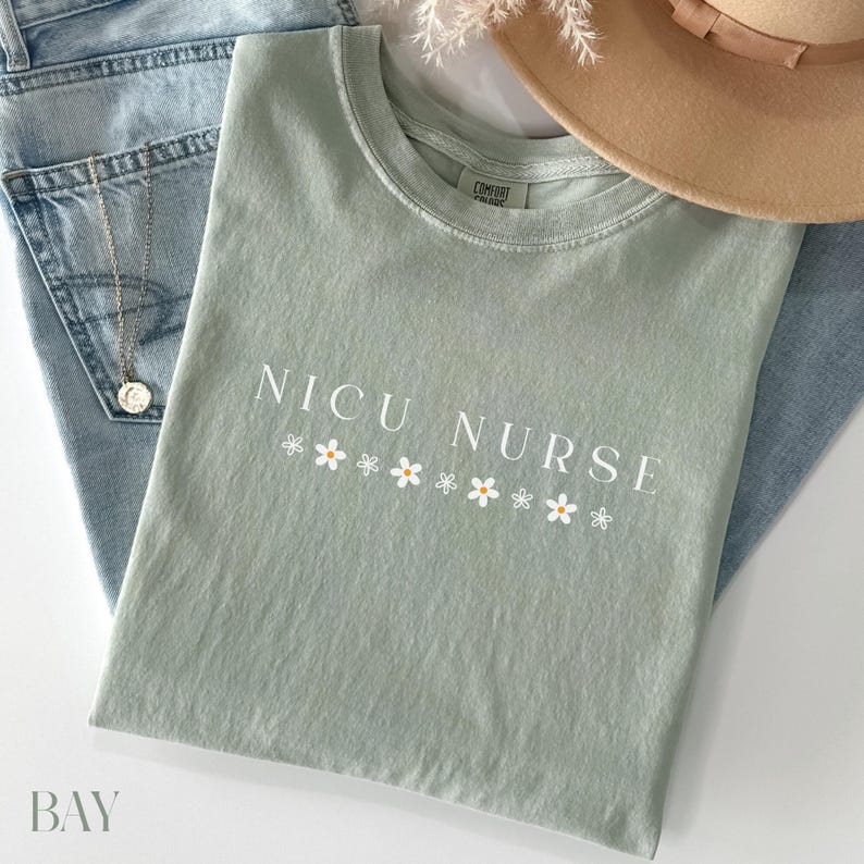 Peut inclure: Un t-shirt vert clair avec le texte "NICU NURSE" en lettres blanches et une rang&eacute;e de petites fleurs blanches avec des centres jaunes sous le texte.