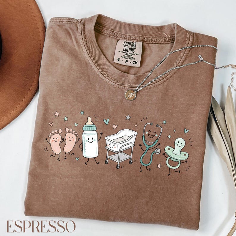 May include: A brown short-sleeve t-shirt with a graphic print featuring a pair of baby feet, a baby bottle, a hospital bassinet, a stethoscope, and a pacifier, all with smiling faces. The shirt has a Comfort Colors brand label at the neck.
