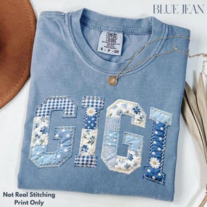 May include: A pale blue Comfort Colors t-shirt featuring the word "GIGI" in a patchwork design. The letters are in various shades of blue with floral and gingham patterns. A gold necklace with a pendant rests on the shirt.