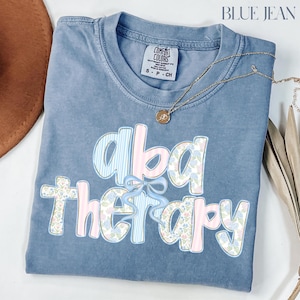 May include: Blue Comfort Colors t-shirt with the words "aba therapy" in a floral and striped design. A gold necklace with a pendant rests on the shirt. The shirt is folded and has a small tag.