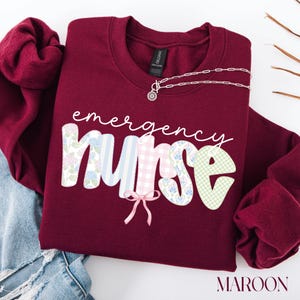 Puede incluir: Sudadera granate con la frase "emergency nurse" en escritura blanca. La palabra "nurse" tiene un dise&ntilde;o colorido. Un collar plateado con un colgante floral est&aacute; sobre la sudadera. La palabra "maroon" est&aacute; en la parte inferior.