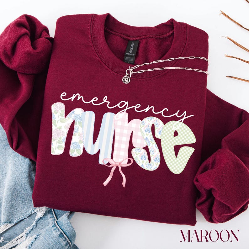 Puede incluir: Sudadera granate con la frase "emergency nurse" en escritura blanca. La palabra "nurse" tiene un dise&ntilde;o colorido. Un collar plateado con un colgante floral est&aacute; sobre la sudadera. La palabra "maroon" est&aacute; en la parte inferior.