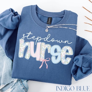 May include: Indigo blue sweatshirt featuring the text "stepdown nurse". The word "nurse" is decorated with floral and striped patterns, and a pink bow. A silver necklace with a sun pendant is on the sweatshirt.