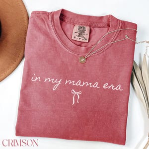 May include: A crimson red t-shirt with the text "in my mama era" in white script with a white bow underneath. The shirt is made by Comfort Colors and has a tag on the front.