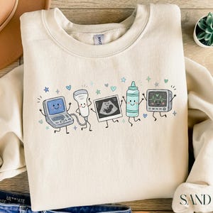 May include: A sand-colored crewneck sweatshirt featuring a graphic print of five anthropomorphic medical items: an ultrasound machine, a transducer probe, a sonogram image, a bottle of gel, and a heart rate monitor, all with smiling faces.